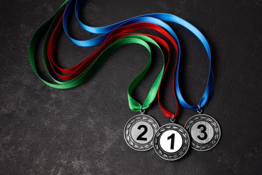 Close-up of three modern silver medals with red, green, and blue ribbons placed on a dark textured surface, showcasing sleek engraved design details.